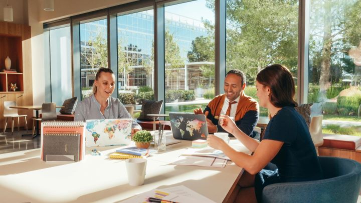 For R at R blog post pexels-kindelmedia-6774432 Three people sit around a table with laptops and other stationery in front of them. Behind them is a big glass window that looks out onto tress and sky. Photograph from Pexels by Kindel Media.