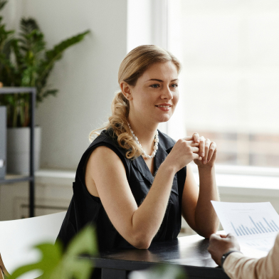getty-images-ZluOgkFWJ4E-unsplash A woman sat at a desk with her elbows propped on the desk, listening to another person. Windows are in the background.