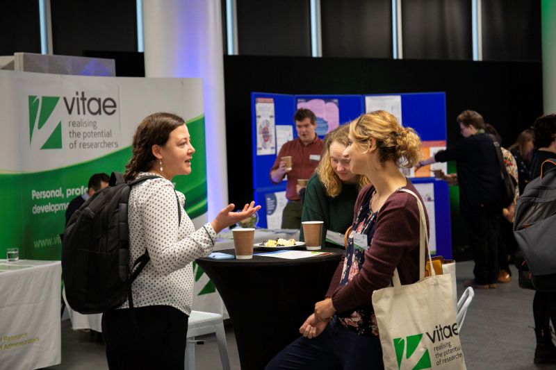 small 0D6A4163 A group of women stand talking in a conference centre with the Vitae logo on a banner behind them.