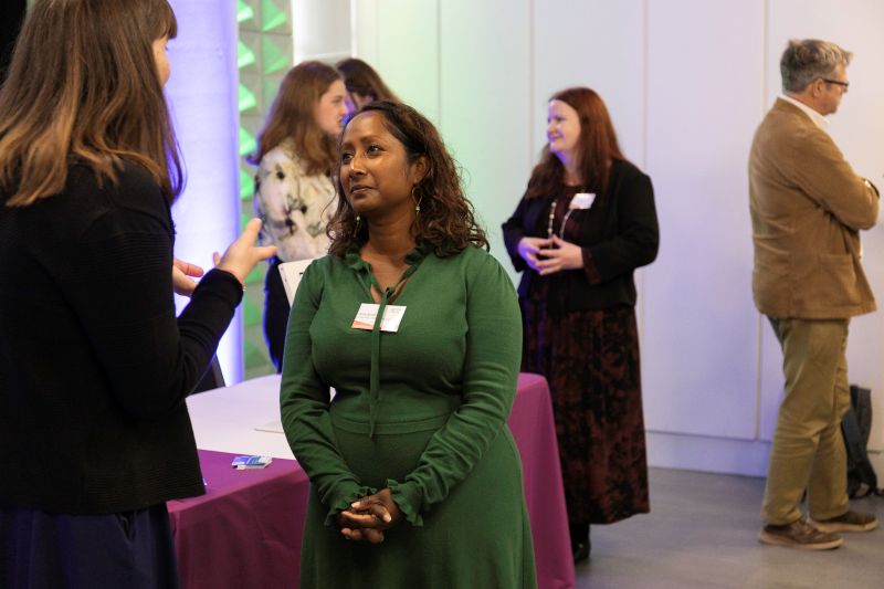 small IMG_6250 A woman wearing a green dress stands and listens to another woman talking, there are various other people talking to each other in the background.