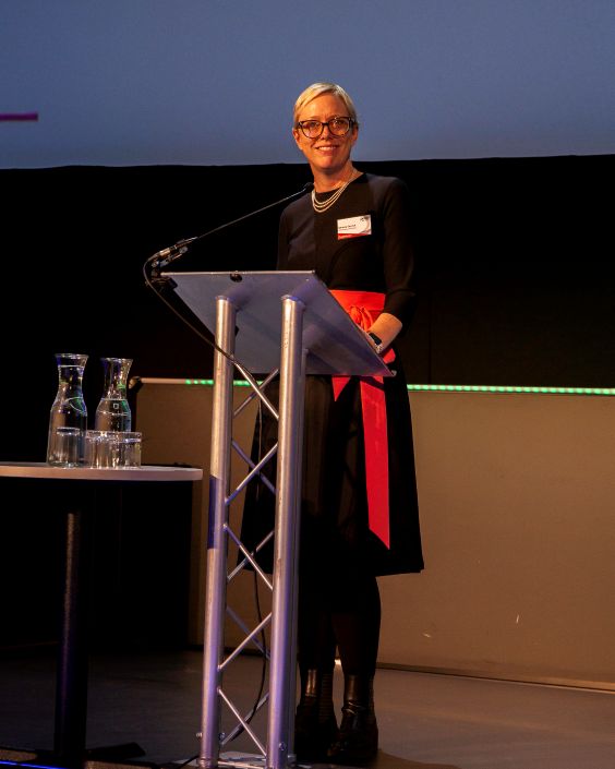 smaller 0D6A4132 A woman wearing a black dress with a red sash stands at a lectern in front of a screen to give a lecture.